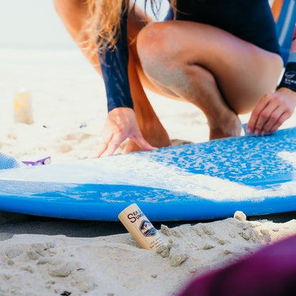 Surfing scenes, the SPF 50 face stick sits in the sand leaning against a woman's surfboard.